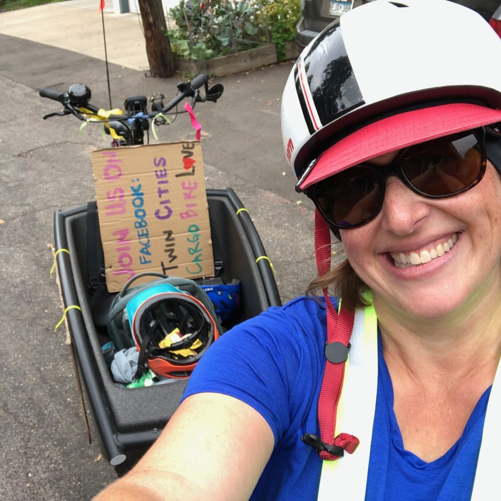 a woman in a bike helmet smiles at the camera with a bakfiets style cargo bike in the background
