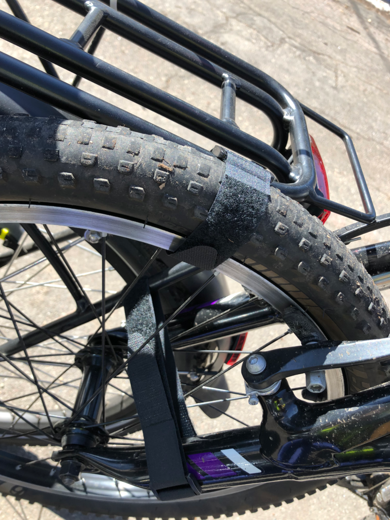 A 20" wheel is strapped to the rear rack of a bakfiets style cargo bike with velcro straps.